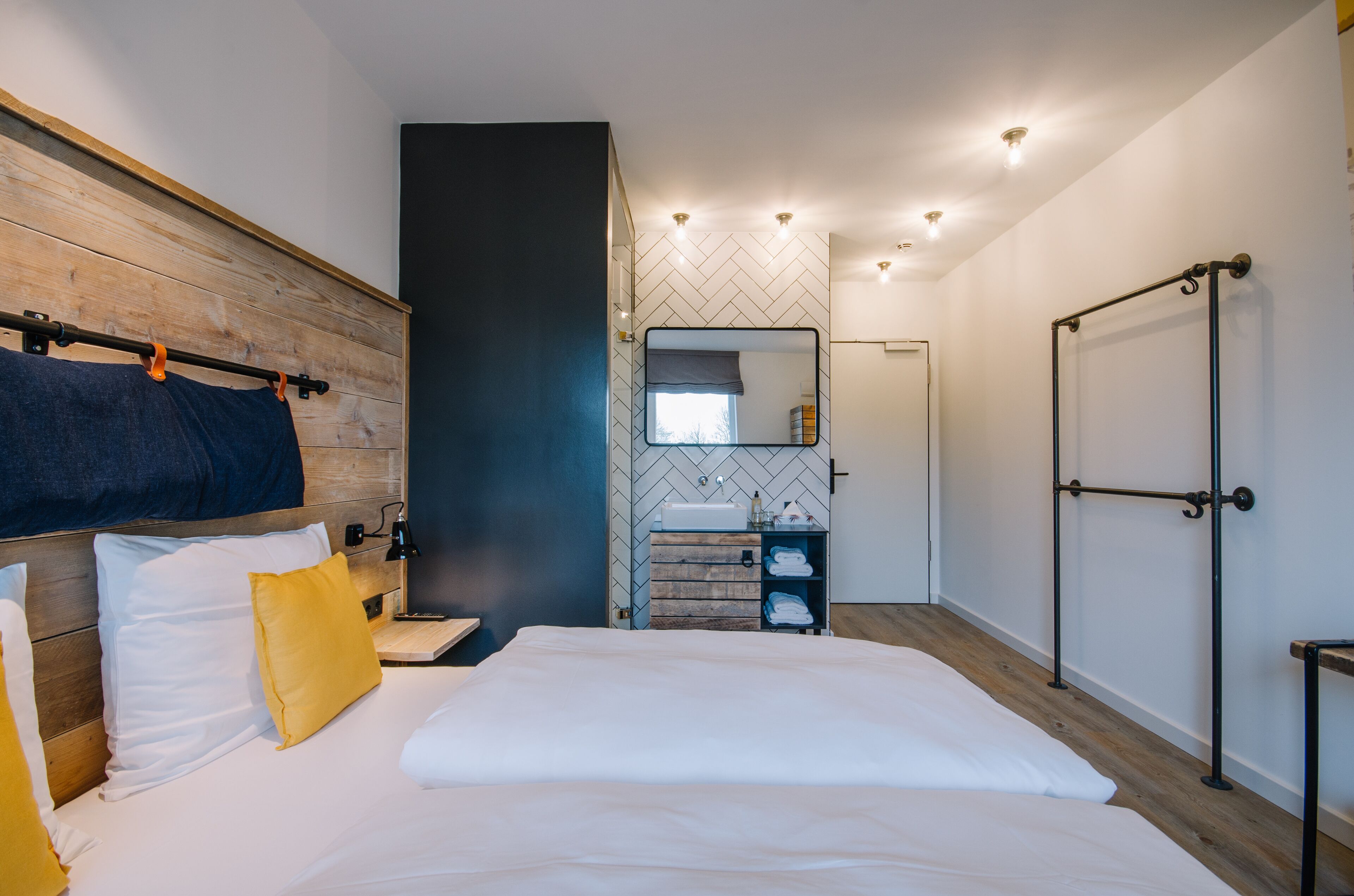 A modern hotel room with a wooden headboard, a large bed with white bedding, a bedside table, a mirror, a built-in bench, a small bathroom with a sink and storage, and a wardrobe with a mirror.