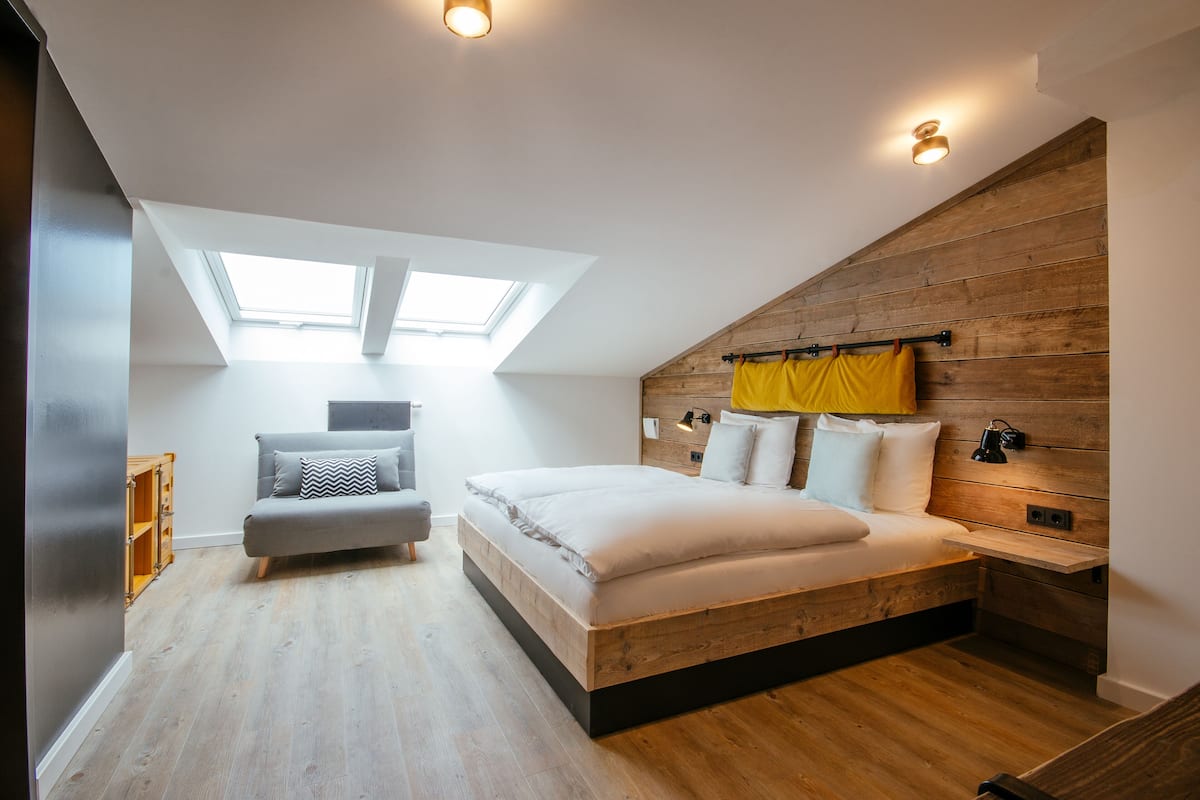 Une chambre avec un plafond en pente, des fenêtres de toit, un lit avec une literie blanche, un canapé gris et un lambris en bois sur les murs.