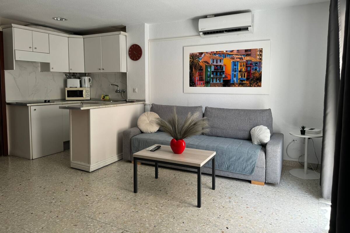 A modern apartment with a kitchenette, a grey sofa, a small table with a vase, and a colorful cityscape painting on the wall.