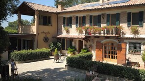 Un edificio de dos pisos con techo de tejas, un balcón con plantas en macetas y un patio con mobiliario exterior.