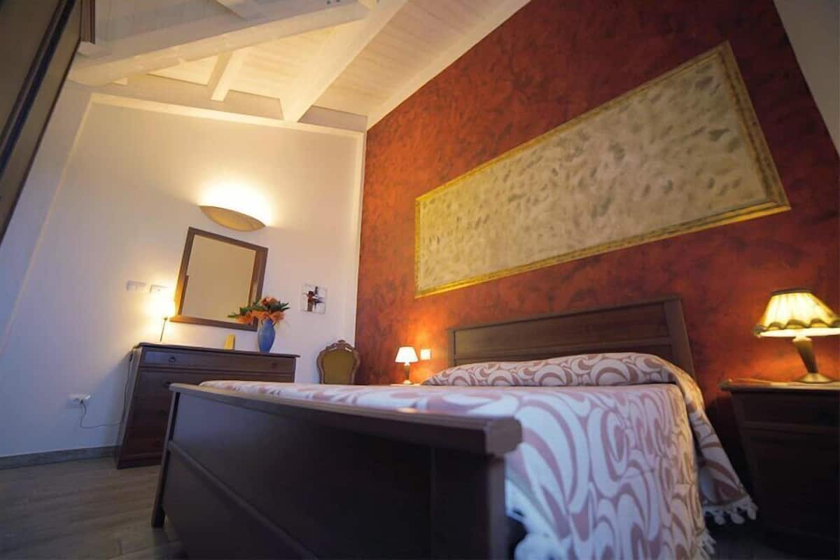 Quarto com cama, luminárias de cabeceira, cômoda, espelho e uma obra de arte na parede.