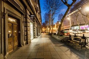 A city street at dusk with buildings on either side, a bench, and a tree-lined sidewalk. - Dat Exx on Freedom Square (Tbilisi)