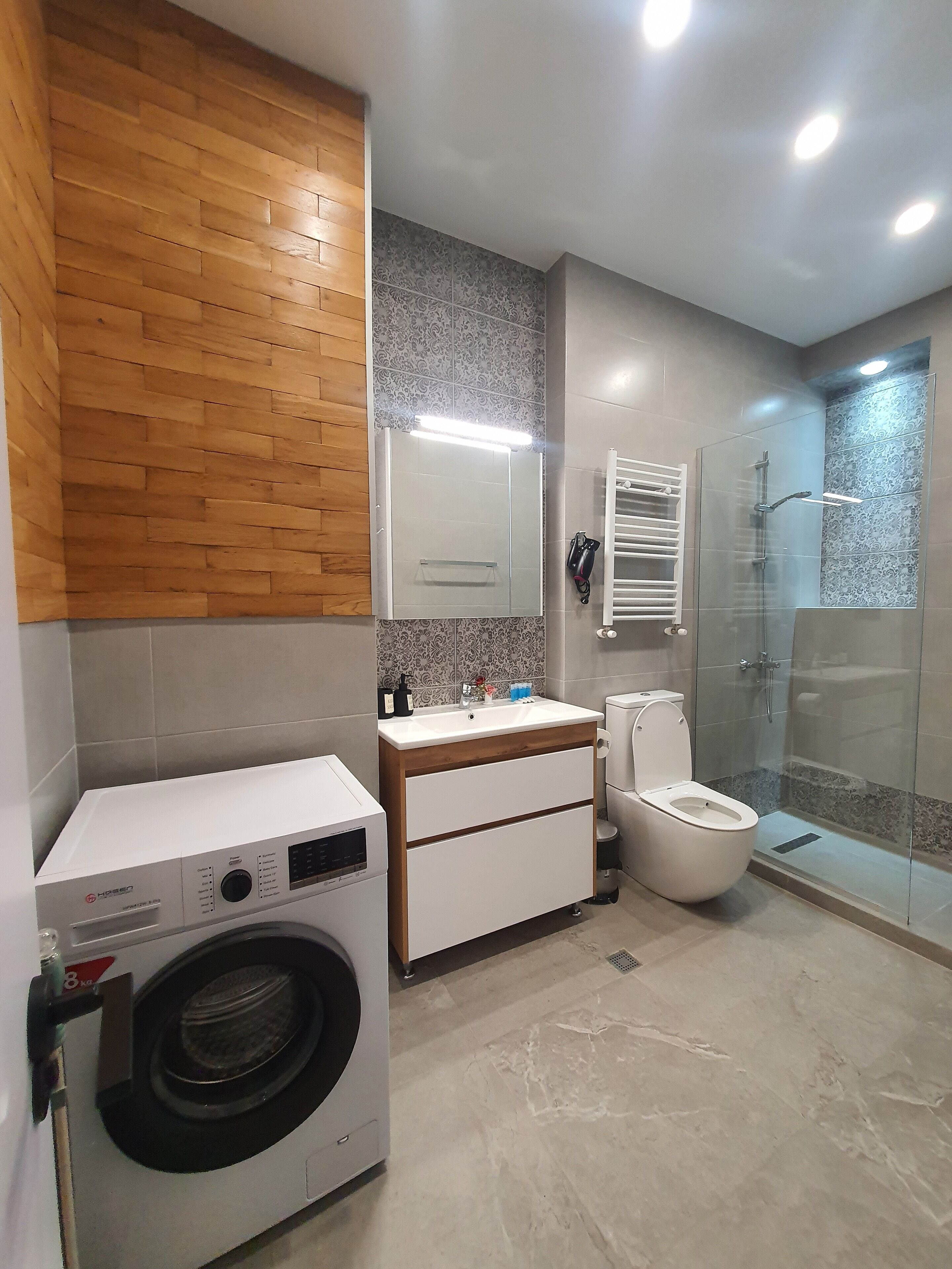 A modern bathroom with a wooden wall, a built-in washing machine, a sink with a mirror, a toilet, and a glass-enclosed shower.