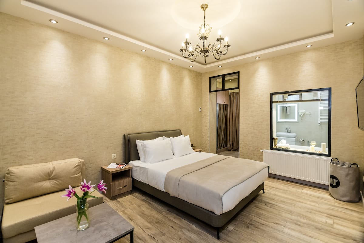 A modern bedroom with a large bed, a sofa, a nightstand, a vase with flowers, and a window with a view of the outside.