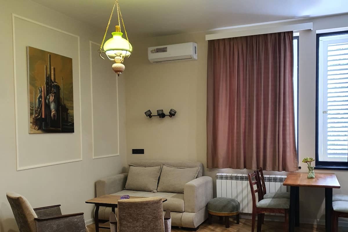 A living room with a sofa, two armchairs, a wooden coffee table, a hanging light fixture, an air conditioning unit, a radiator, and a window with curtains.