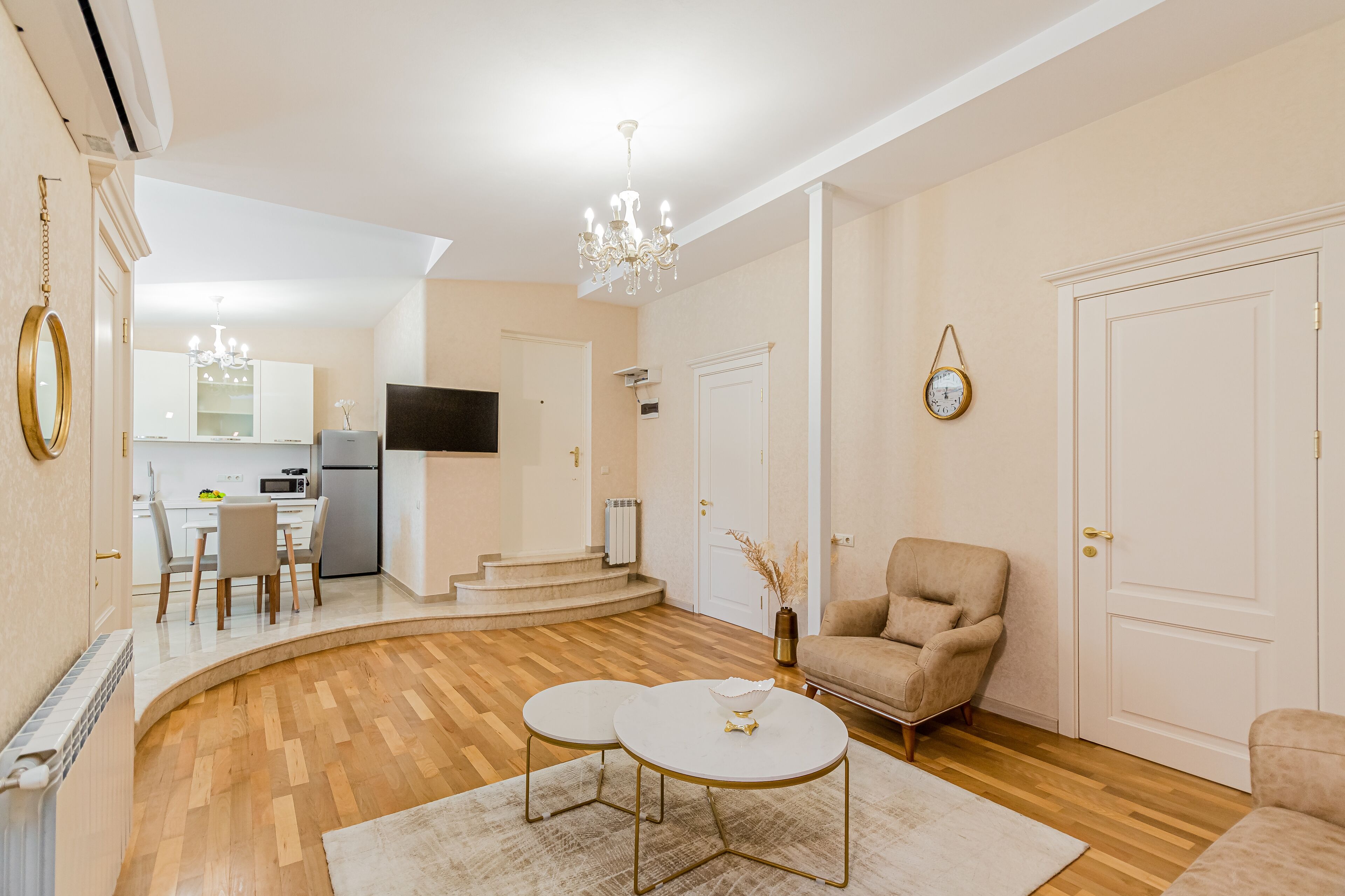 Una sala moderna con piso de madera, una araña y un sillón color crema, además de un comedor con una mesa y sillas blancas.
