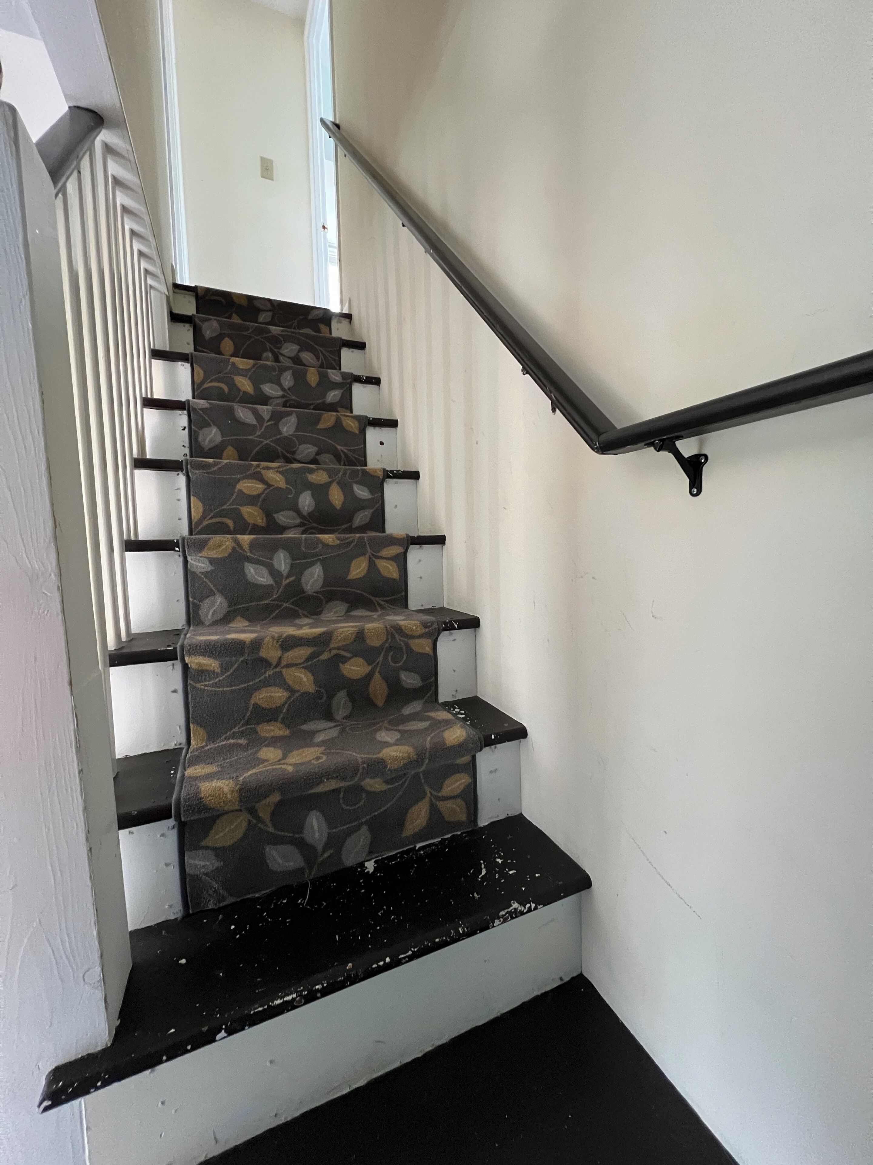 A staircase with patterned carpet and black risers, leading to a landing with a white wall and a metal handrail.