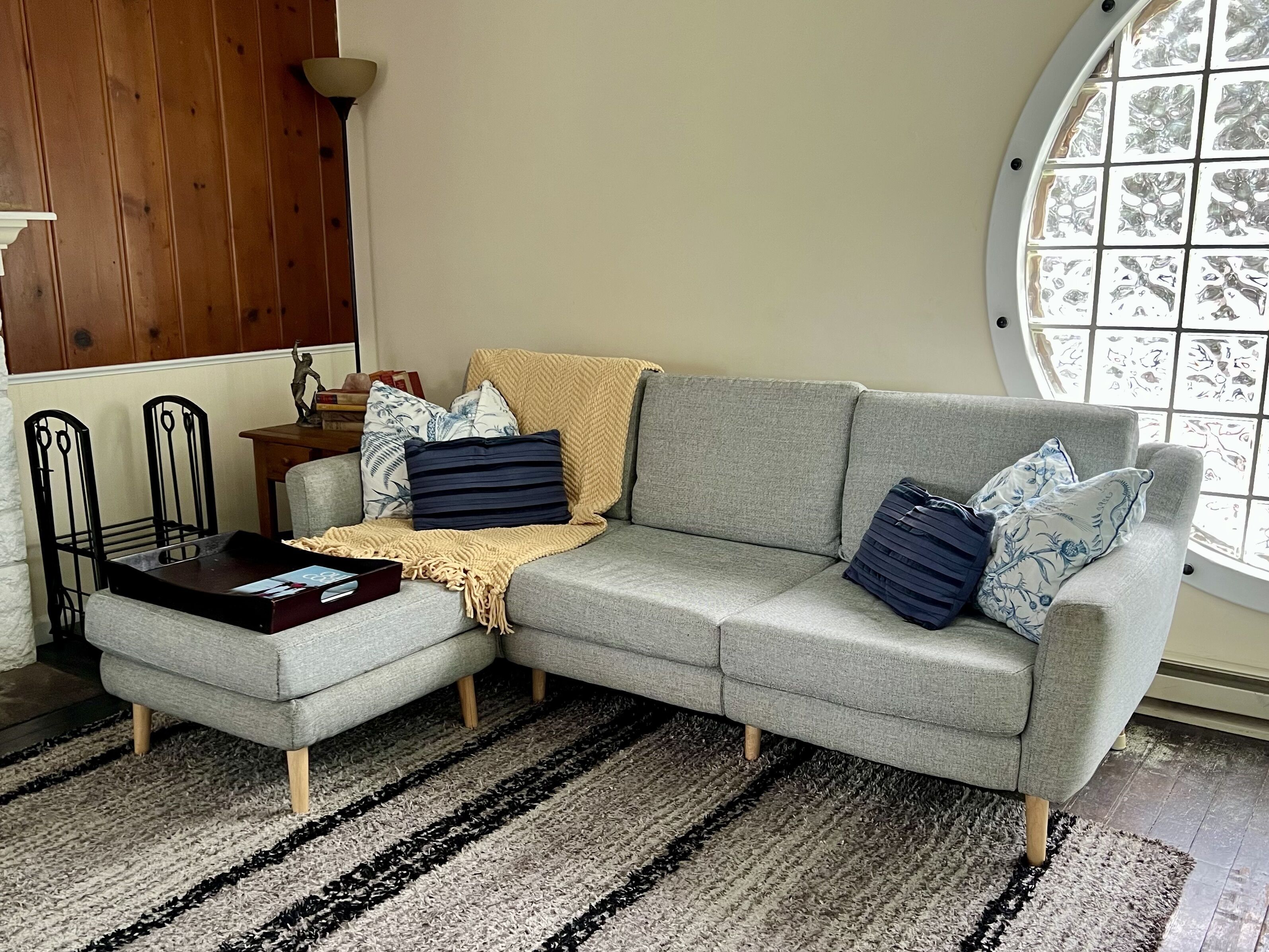 A grey sectional sofa with a beige throw blanket, a small ottoman, and multiple pillows. A round window with a grid pattern is visible in the background.