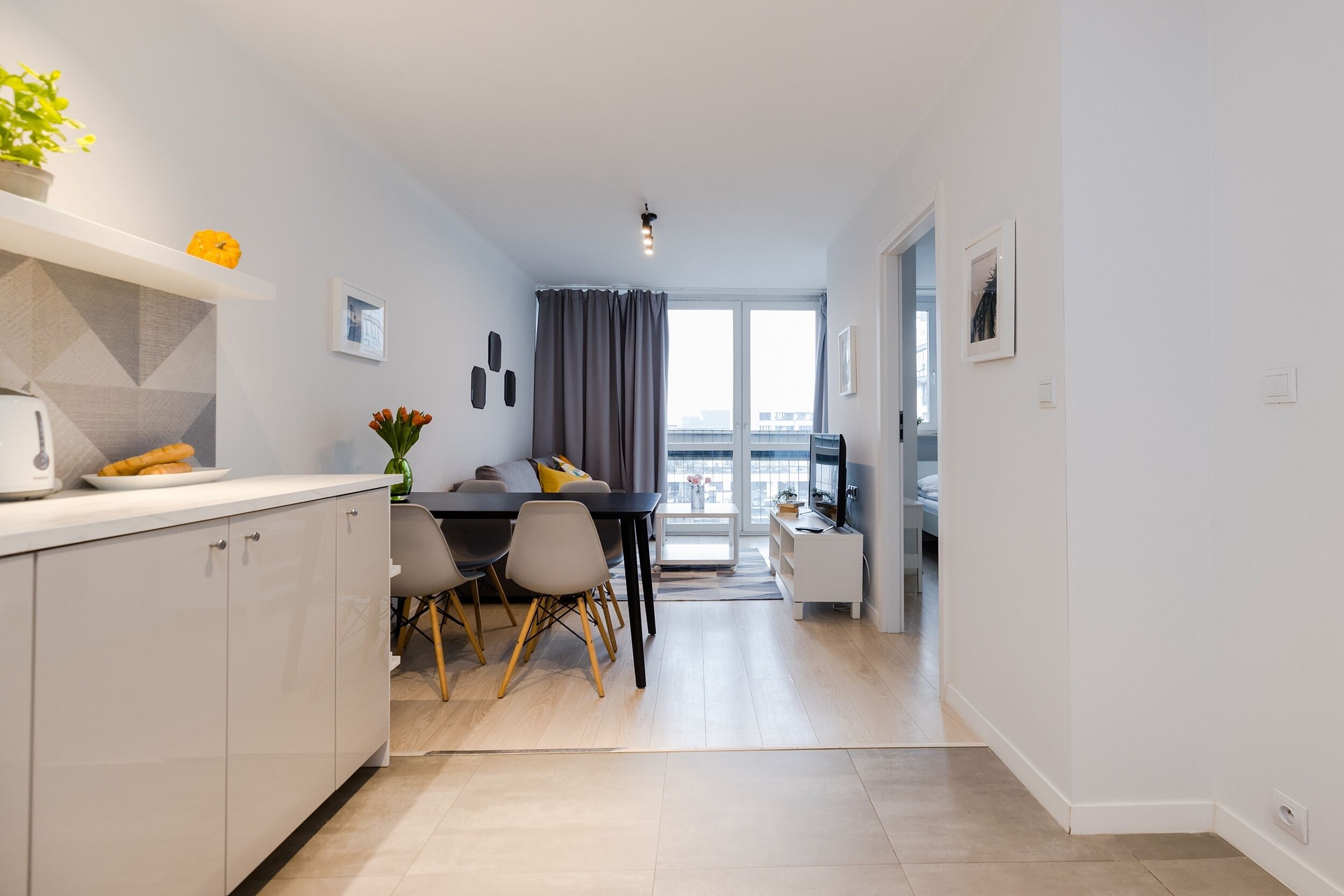A modern apartment with a kitchen, dining area, and living space. It features a large window, a flat-screen TV, and a view of the city.
