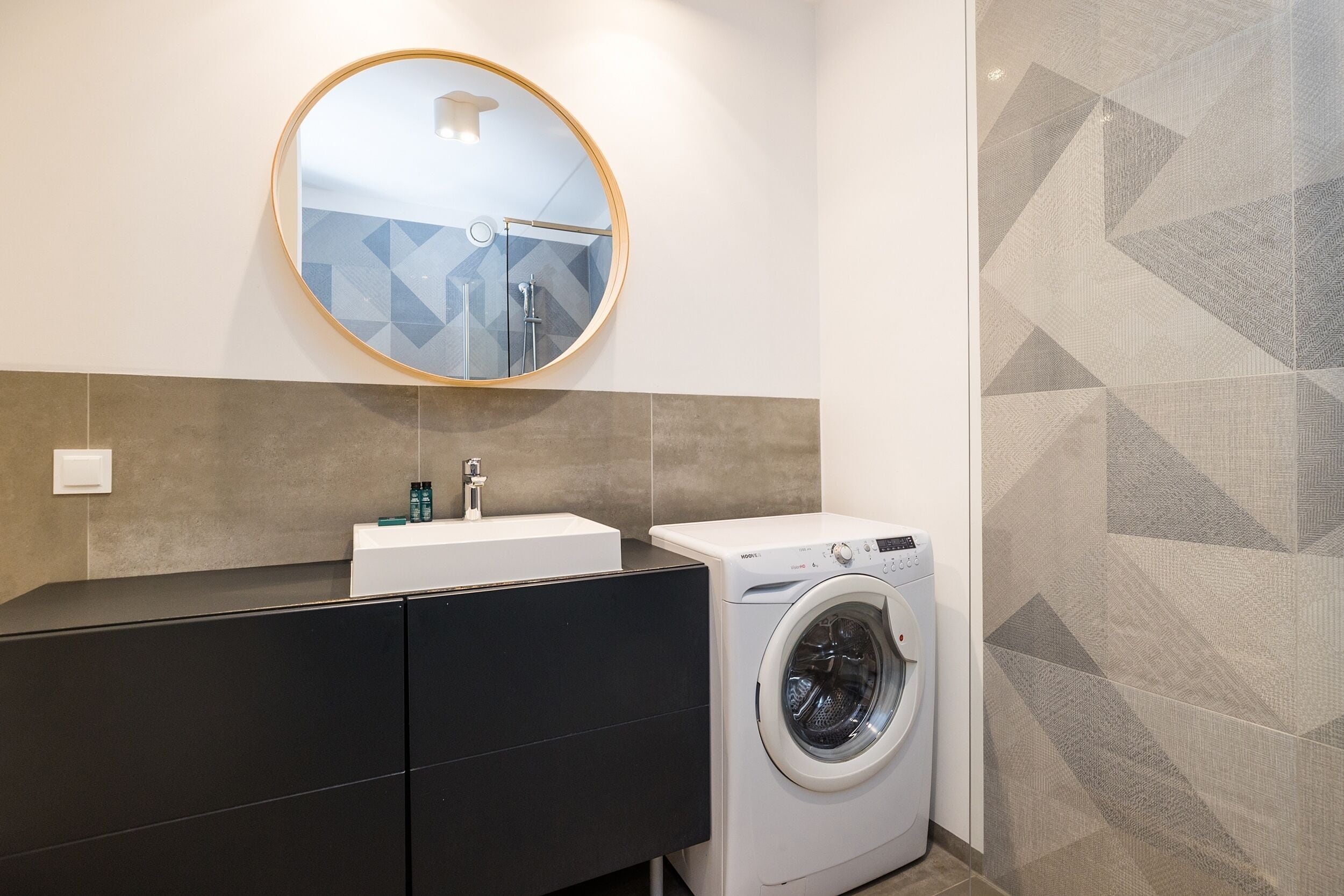 A modern bathroom with a round mirror, a washing machine, and a dark-colored vanity.