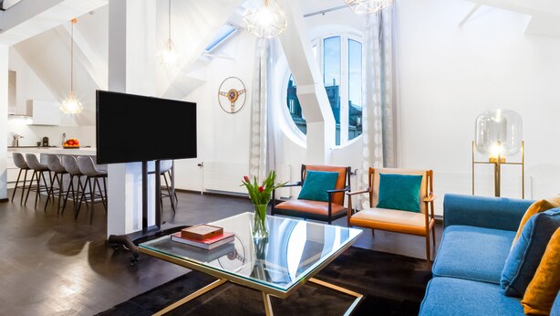 Ein modernes Wohnzimmer mit einem Glastisch, einem Flachbildfernseher und einer Couch. Zwei Sessel mit Kissen, ein Essbereich mit Stühlen und ein großes Fenster mit Vorhängen sind ebenfalls vorhanden.