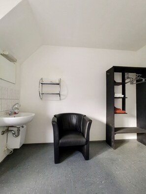 Un baño con un lavamanos blanco, una silla negra y un estante negro con zapatos.
