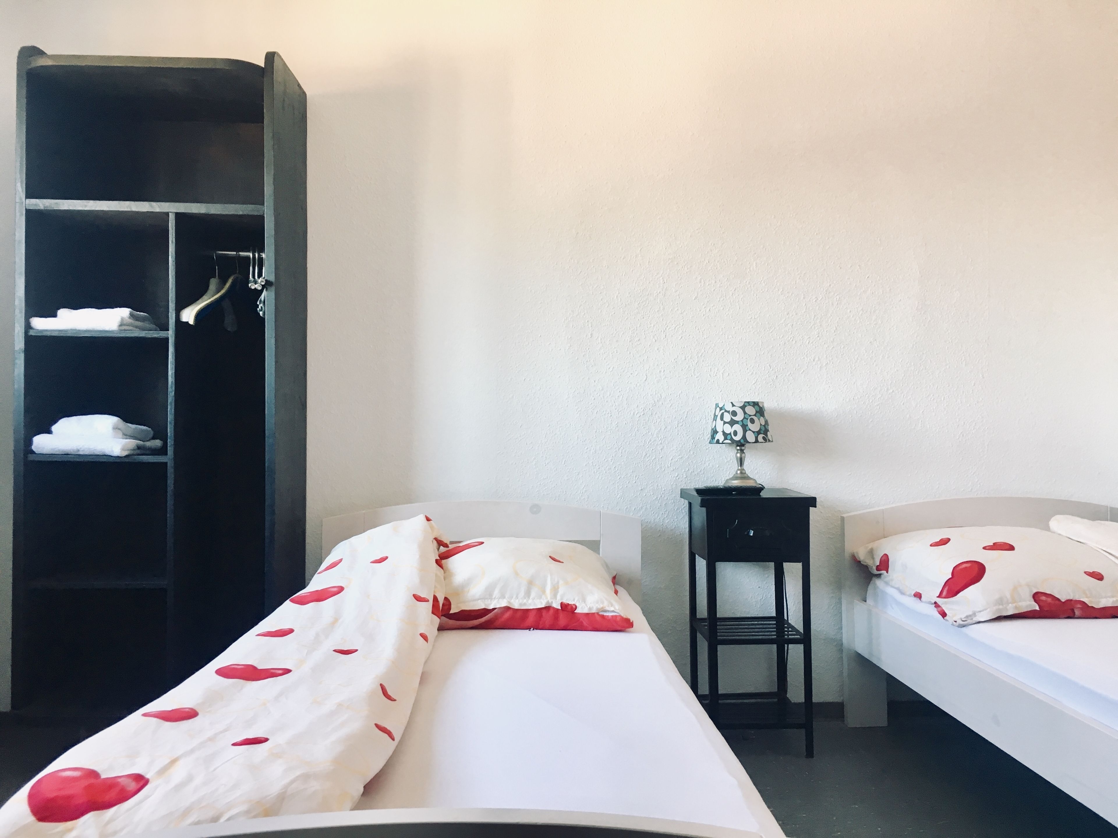 A room with two beds, a wardrobe, and a small table with a lamp.