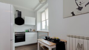 A modern kitchen with white cabinetry, a black hanging pot, a wooden table, and a window with blinds. - ShortStayPoland Staszica B76 (Warsaw)