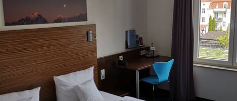 Quarto de hotel com duas camas de solteiro, uma mesa de trabalho com cadeira, um quadro na parede e uma janela com vista para edifícios.