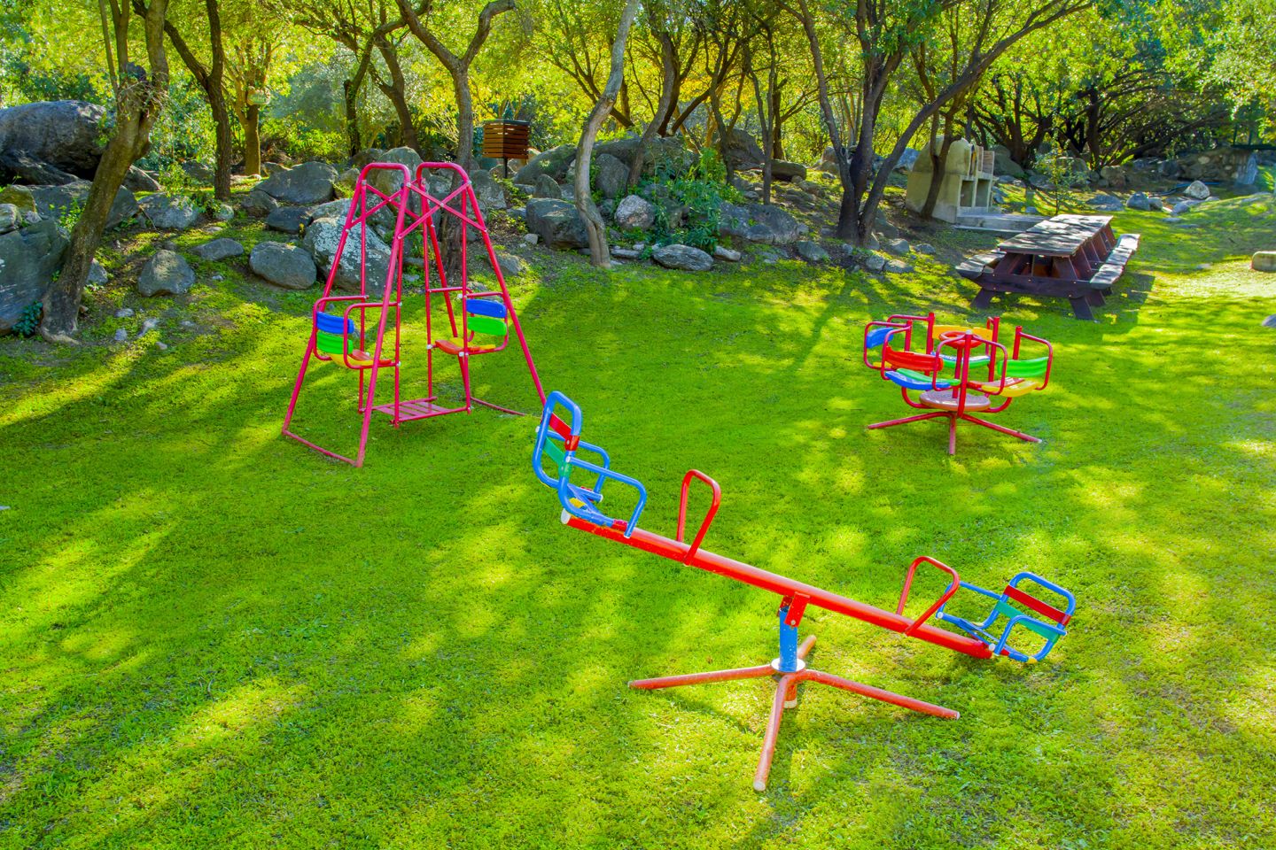 A playground with swings and a seesaw in a grassy area surrounded by trees.
