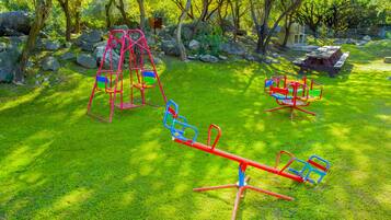 A playground with swings and a seesaw in a grassy area surrounded by trees.