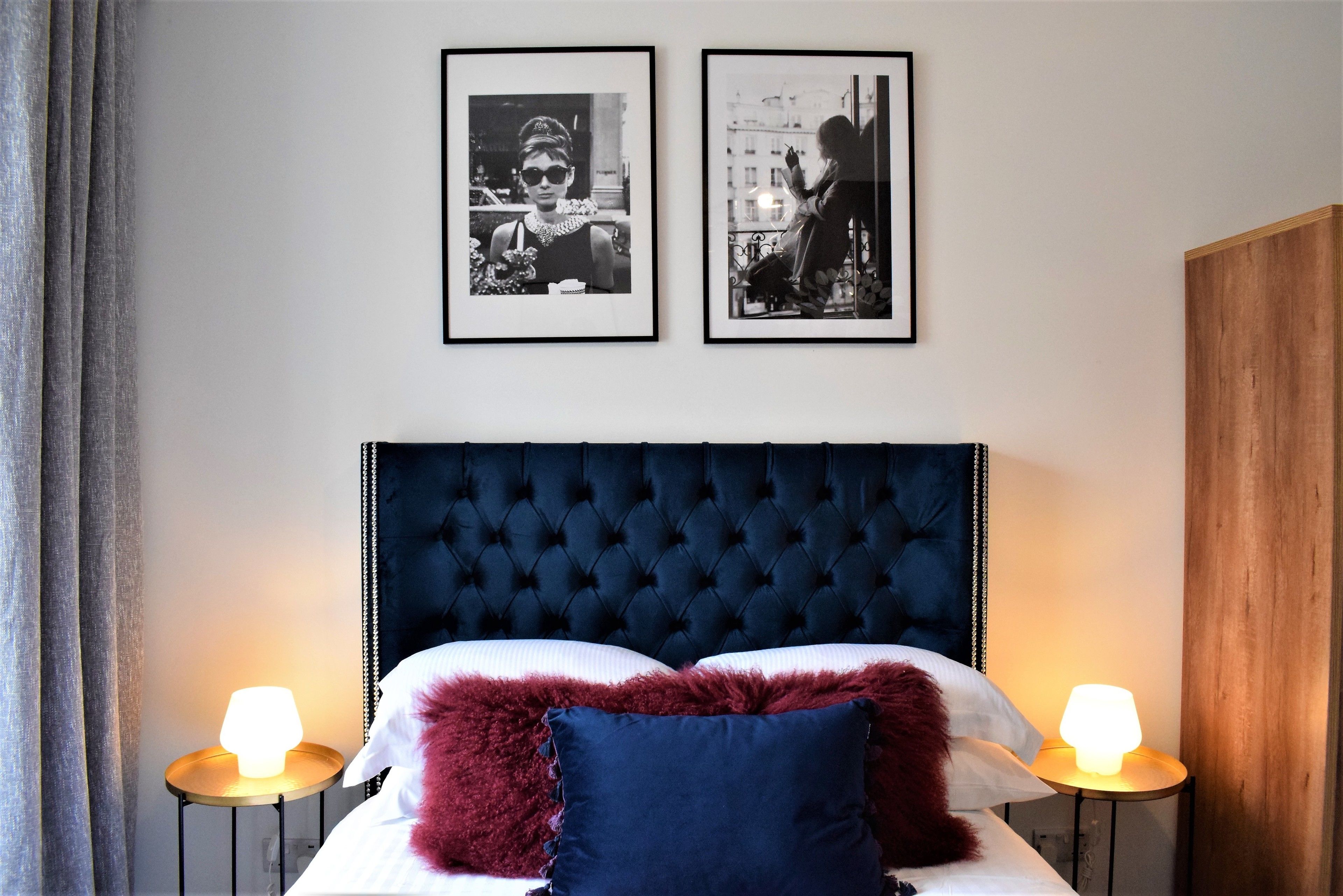 A bedroom with a tufted headboard, two bedside lamps, a bed with white and red pillows, and two framed photographs on the wall.