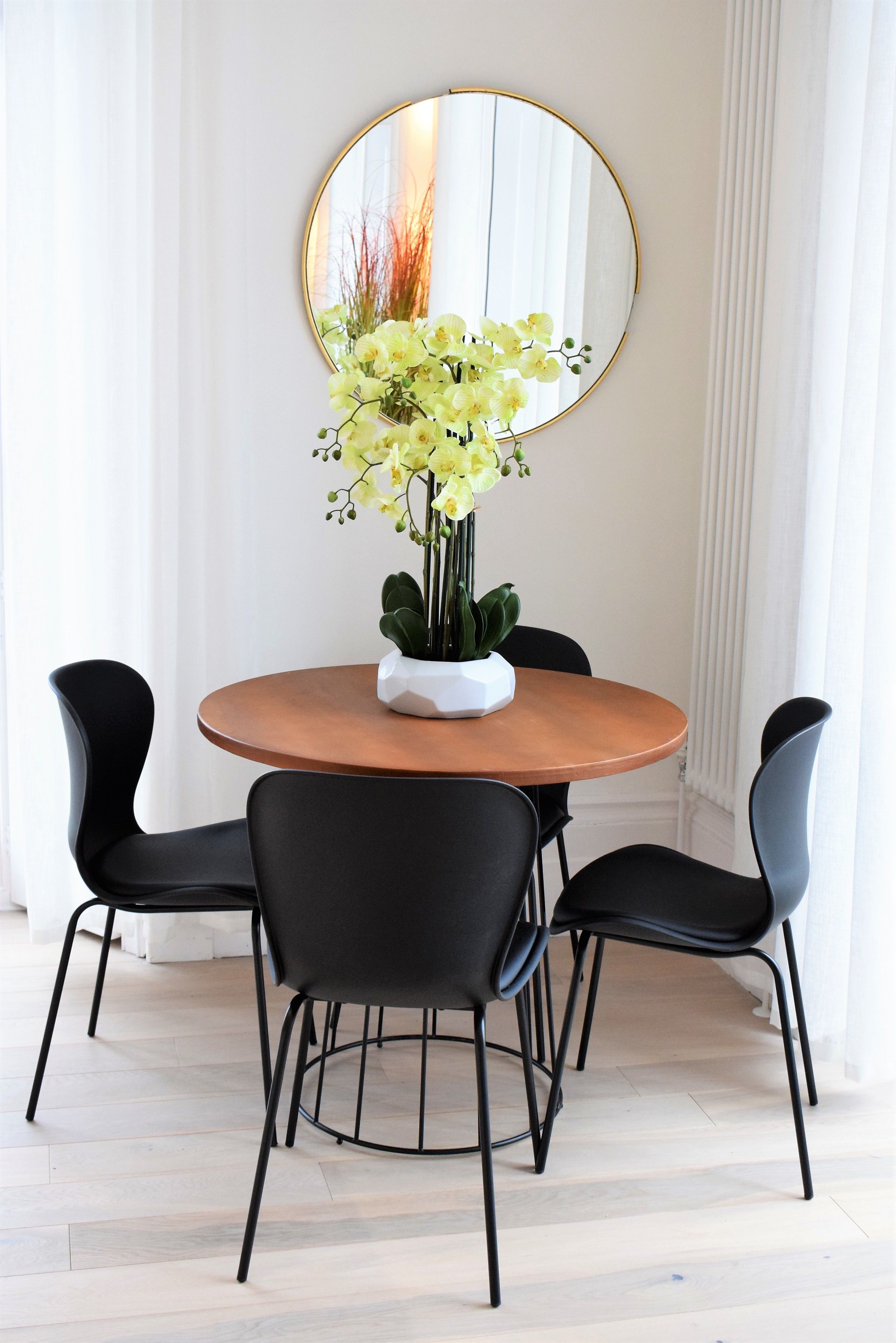 A dining area with a round wooden table, four black chairs, and a white vase with yellow flowers.