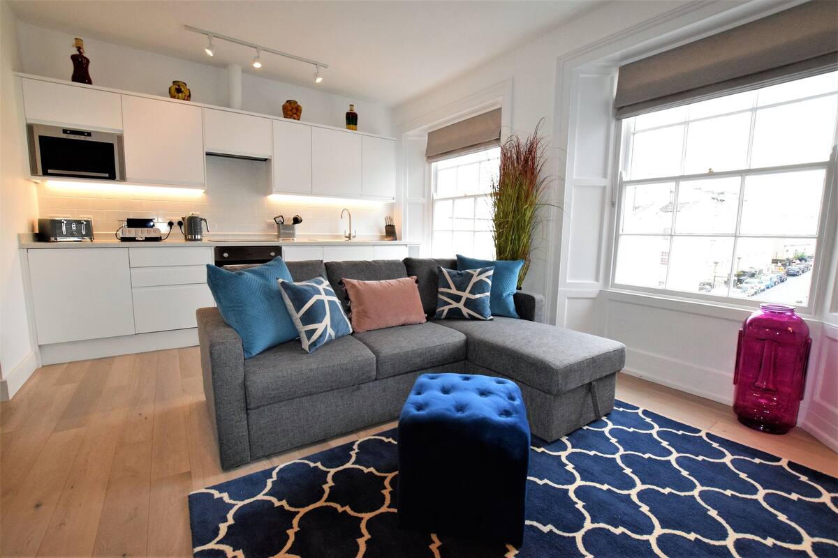 A modern living room with a grey sectional sofa, a blue ottoman, and a patterned rug.