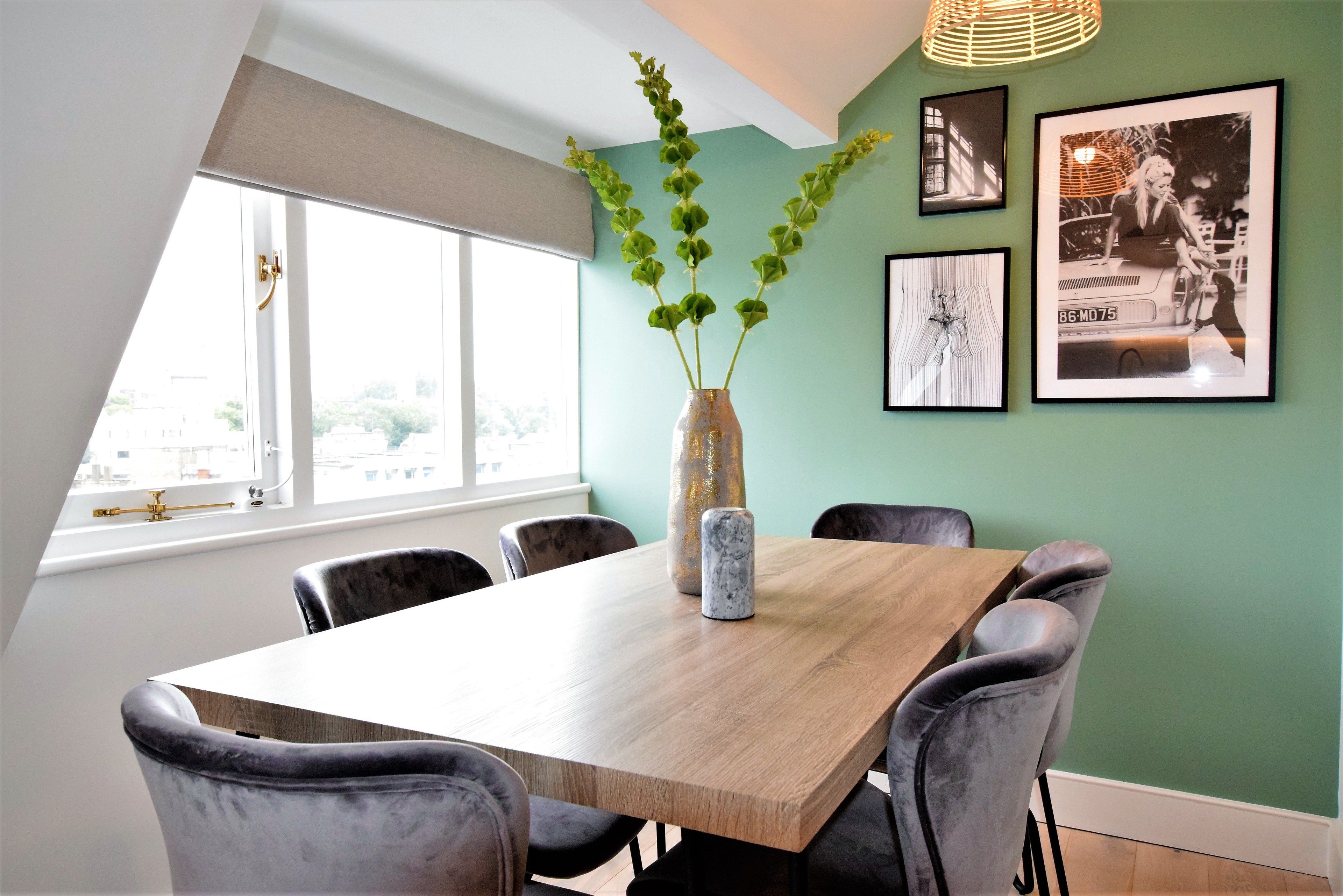 A dining area with a wooden table, grey upholstered chairs, a vase with green plants, and framed pictures on the wall.