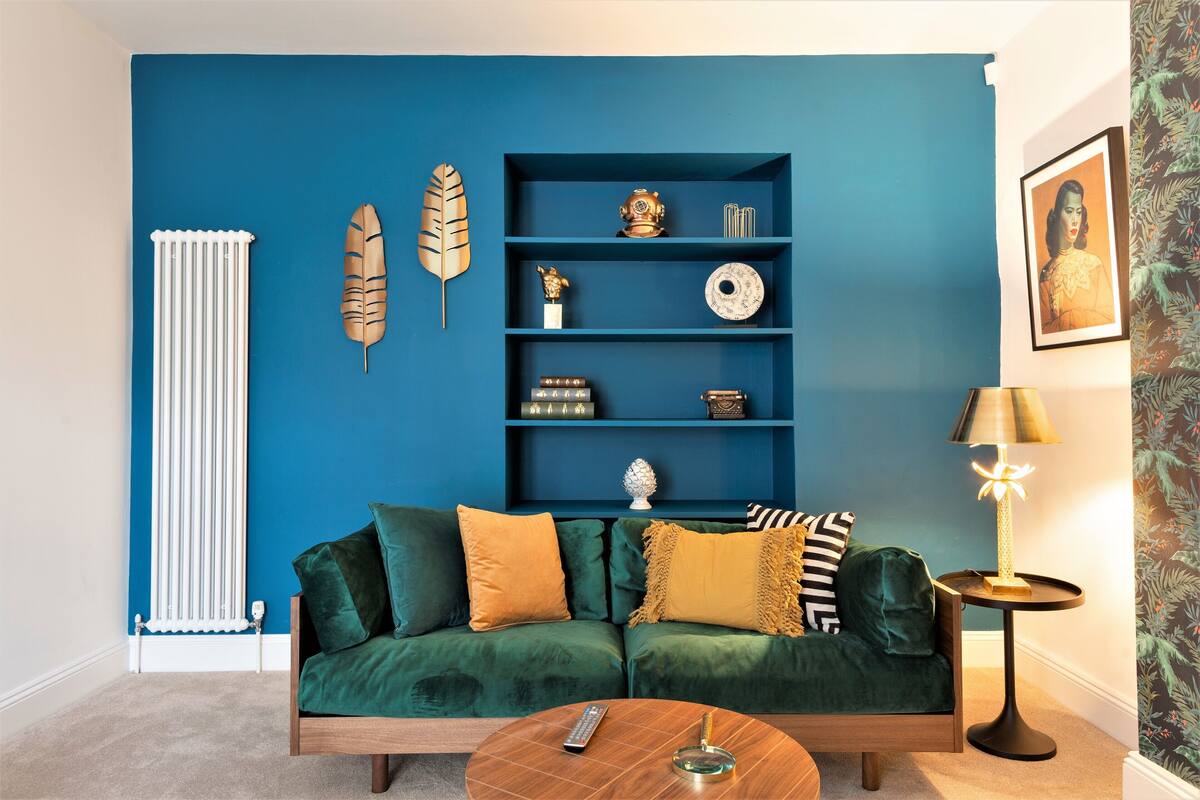 A room with a blue accent wall, a green sofa with yellow and black cushions, a wooden coffee table, and a side table with a lamp.