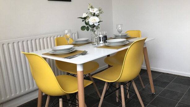 A dining table set with plates, cutlery, and glasses, surrounded by yellow chairs, with a radiator and a framed quote on the wall. - Swale Corner (Wellingborough)