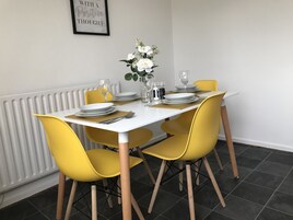 Una mesa de comedor dispuesta con platos, cubiertos y vasos, rodeada de sillas amarillas, con un radiador y una cita enmarcada en la pared.