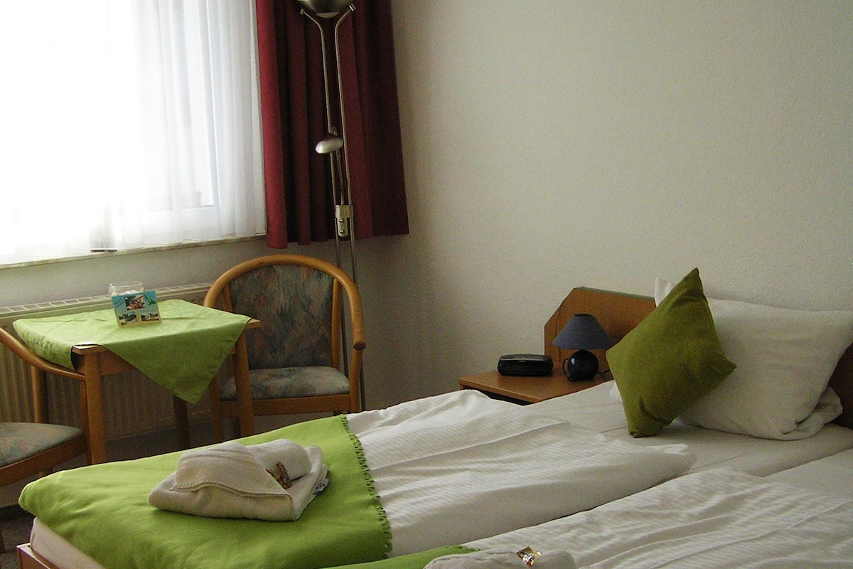 A hotel room with two single beds, a small table with a chair, a lamp, and a window with curtains.