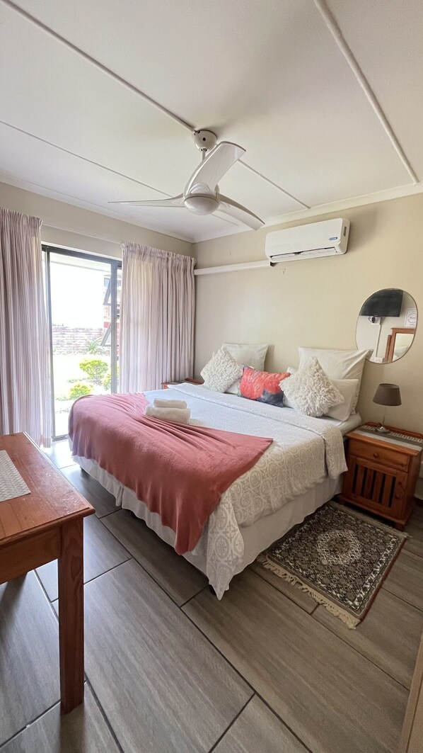 A hotel room with a bed, a ceiling fan, an air conditioning unit, a wooden nightstand, a small table, and a view through a window with curtains.