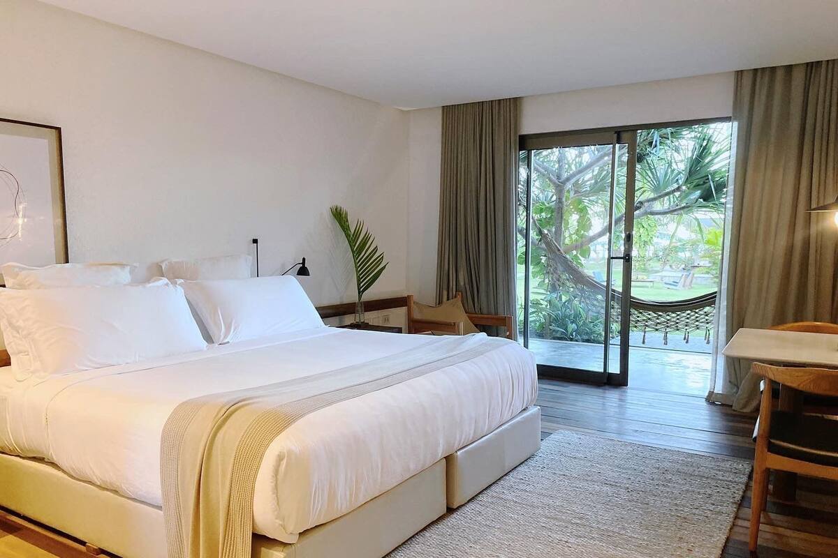 Une chambre spacieuse avec un grand lit, un bureau et une vue sur un jardin luxuriant, accessible par une porte-fenêtre coulissante.