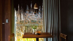 A wooden table with two chairs, a hanging light fixture, and a view of a stone wall and greenery through a window with curtains. - Paru Boutique Hotel (Passo de Camaragibe)