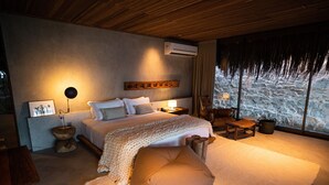 A bedroom with a large bed, a bedside table, a chair, a wooden headboard, and a view of a rocky landscape through the window. - Paru Boutique Hotel (Passo de Camaragibe)