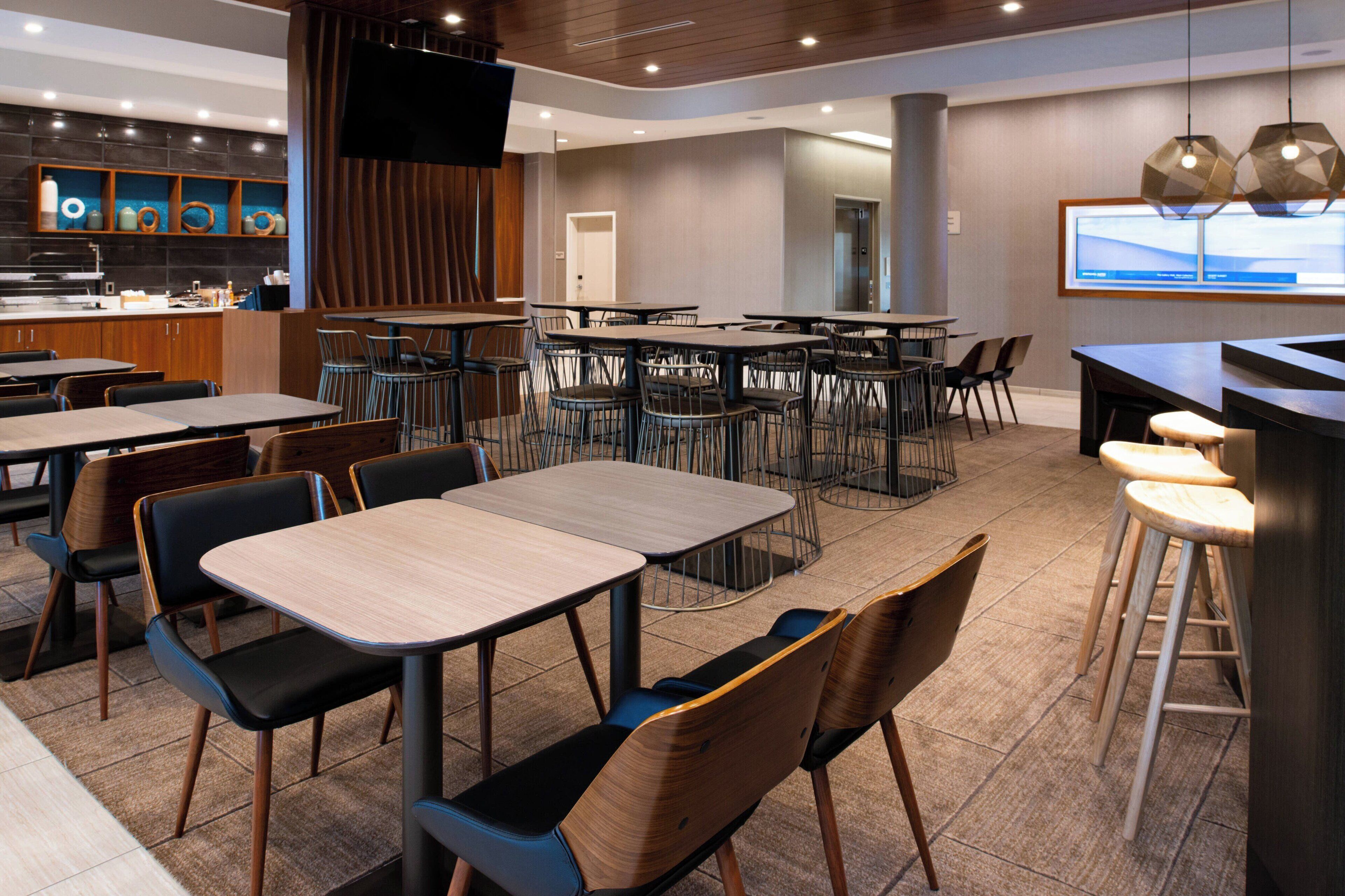 A modern restaurant with multiple dining tables and chairs, a bar area, and a television screen.