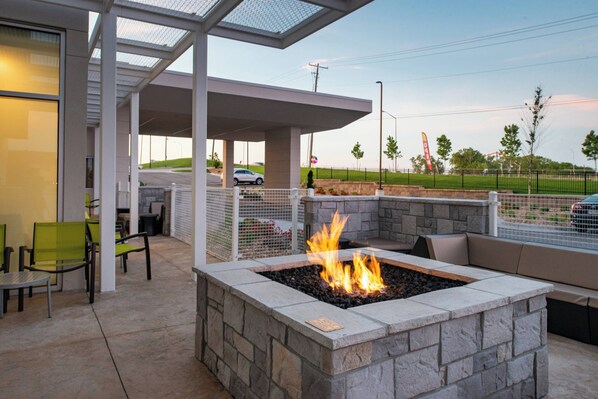 An outdoor patio with a stone fire pit and seating, a covered area with chairs, and a view of a parking lot and distant landscape.