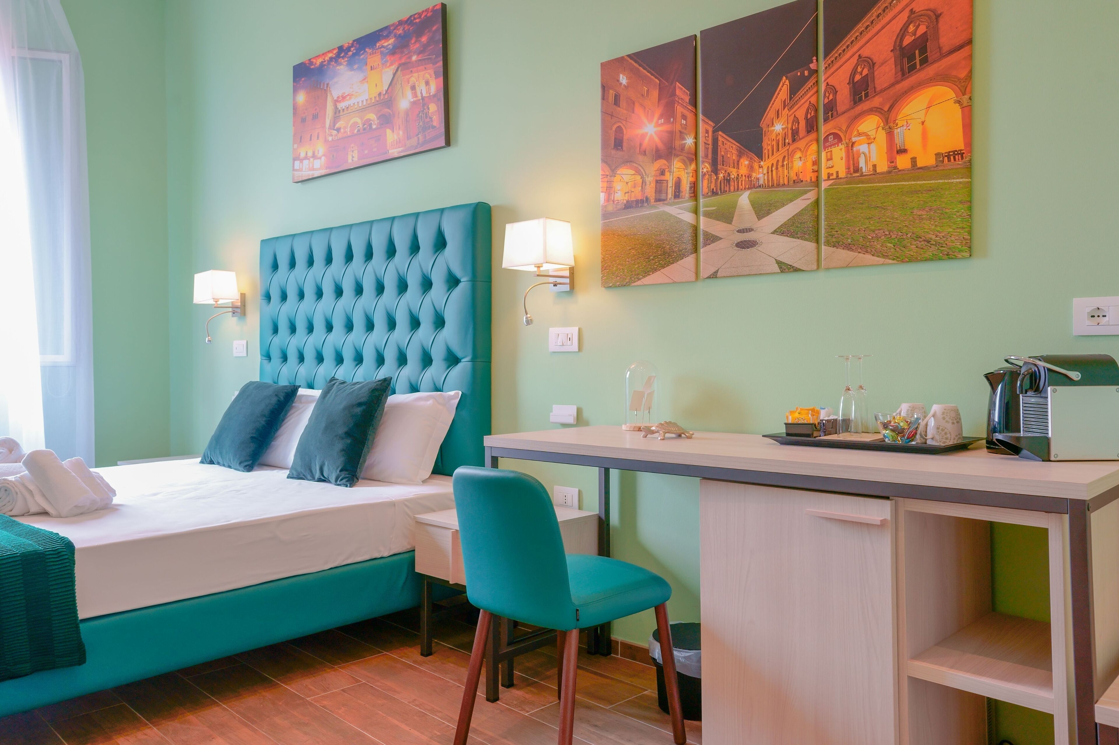 A hotel room with a bed, a desk, and two chairs. There are paintings on the wall and a lamp on the desk.