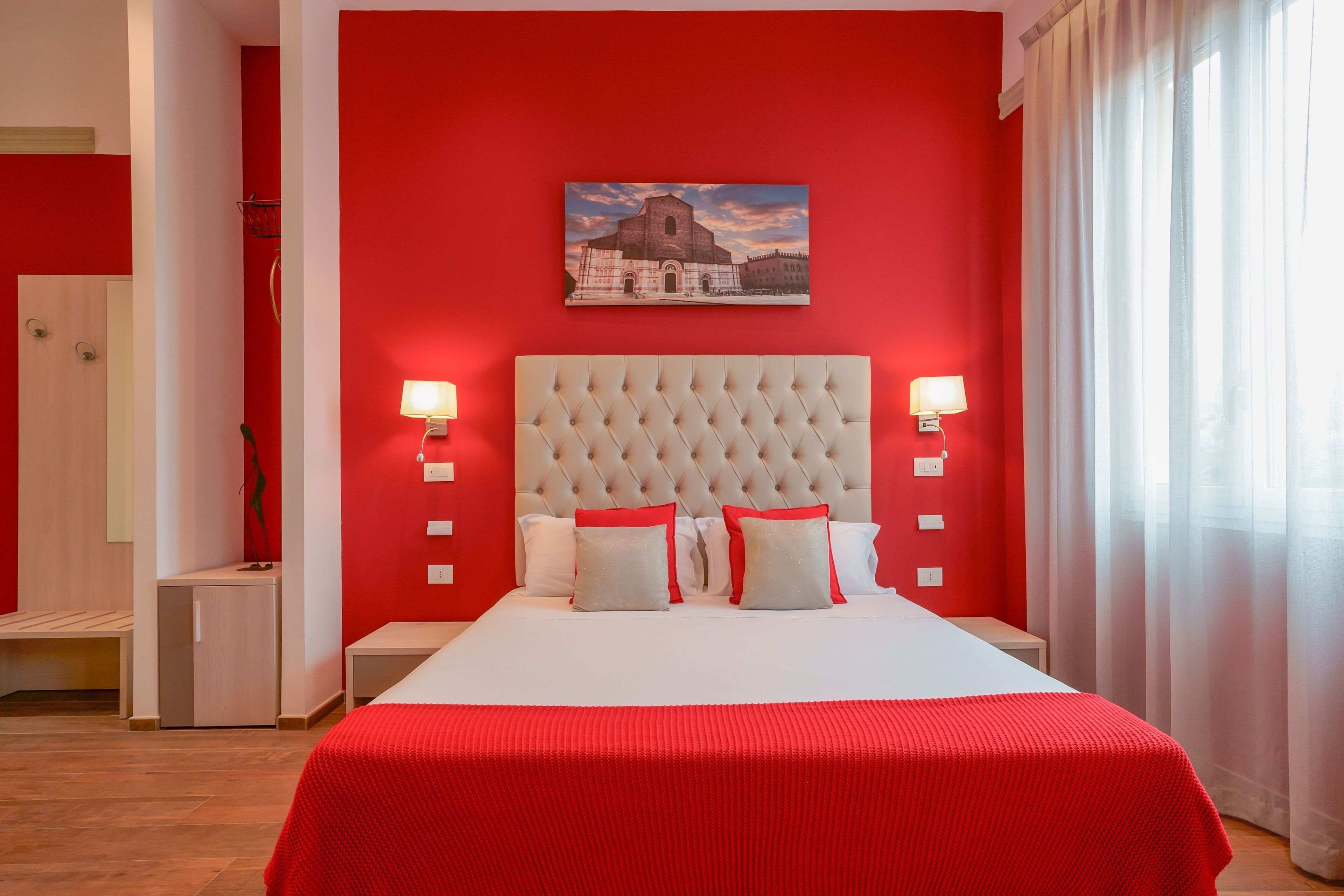 Quarto com parede vermelha, uma cama grande com roupa de cama branca e vermelha, duas luminárias de cabeceira e uma pintura de um edifício.