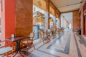 Un café interior con sillas y mesas de mimbre, piso de baldosas y vista a la calle a través de grandes ventanales.