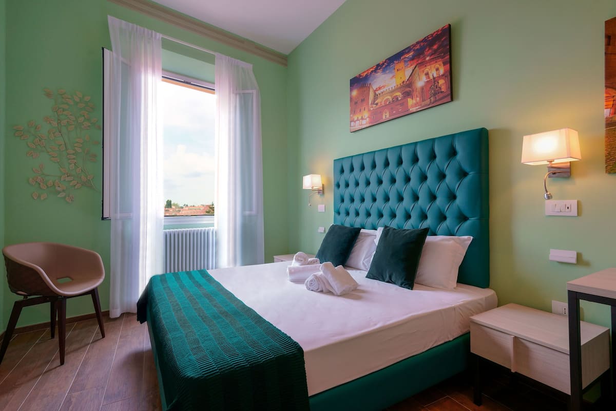 A hotel room with a large bed, a chair, a table, and a view of the sea through the window.