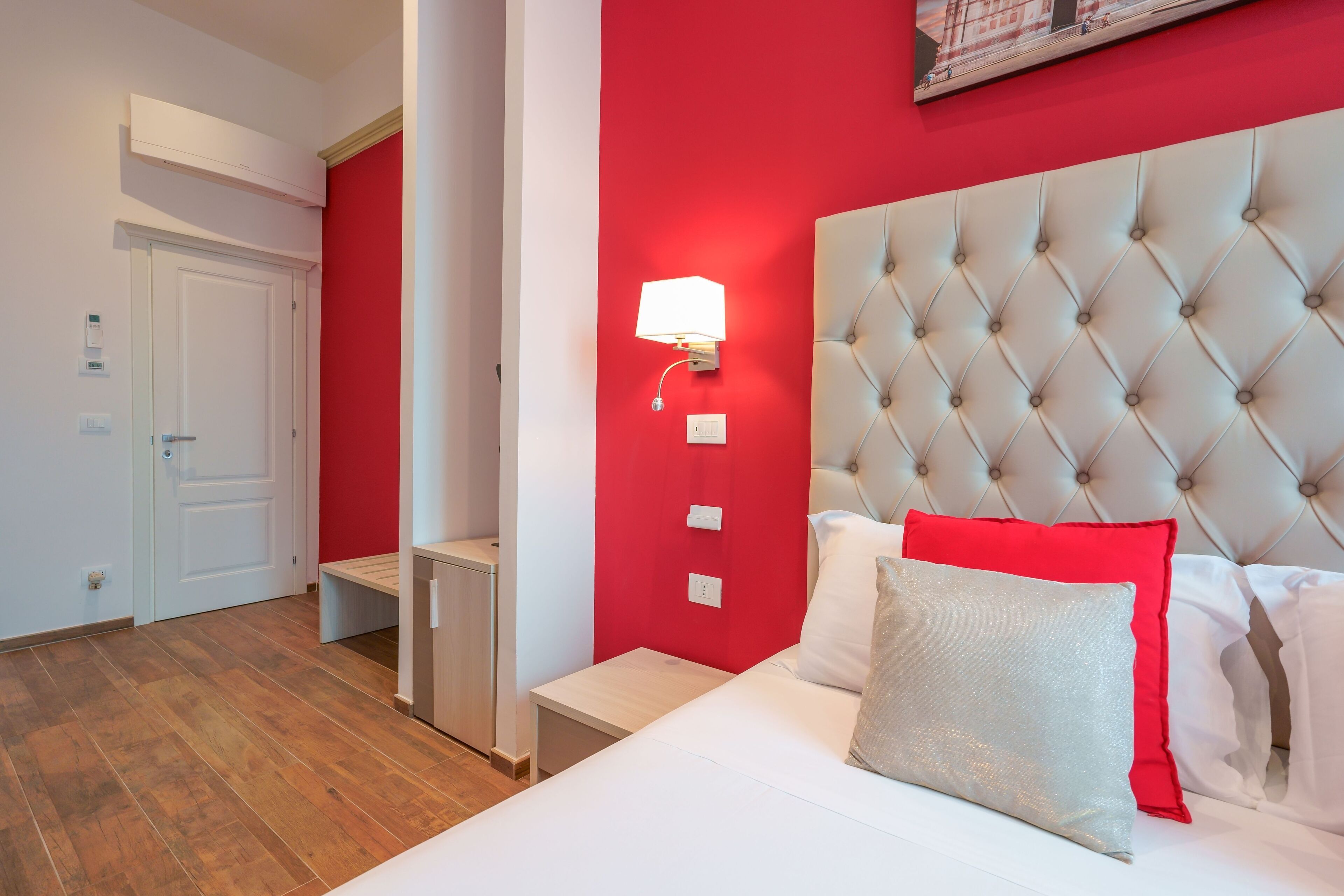 Une chambre d’hôtel avec un mur d’accent rouge, une tête de lit capitonnée, un lit blanc avec des coussins rouges et argentés, une table de chevet et une petite table de nuit avec une lampe.