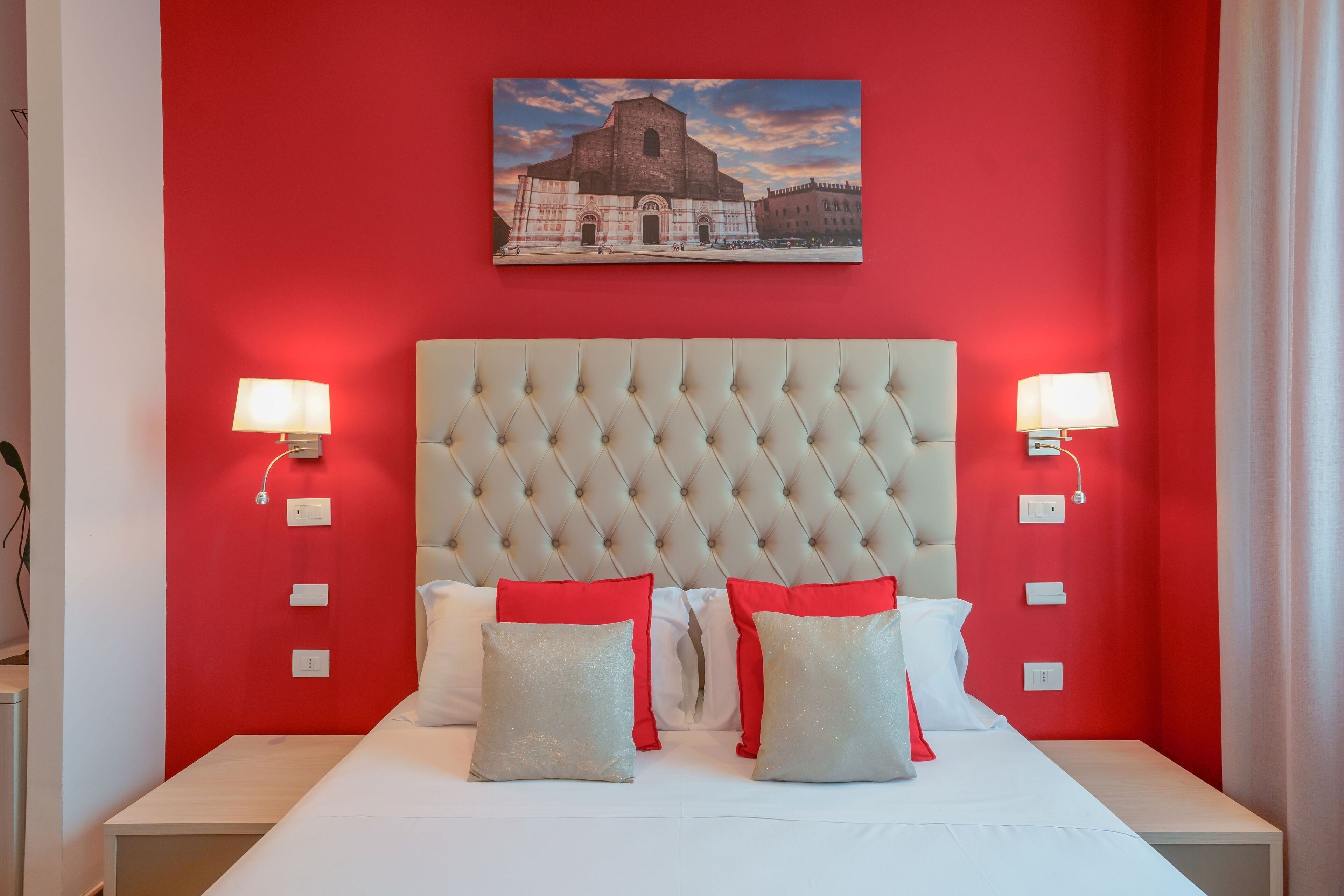 Quarto de hotel com parede vermelha, cabeceira estofada, duas luminárias de cabeceira e um quadro de um edifício.