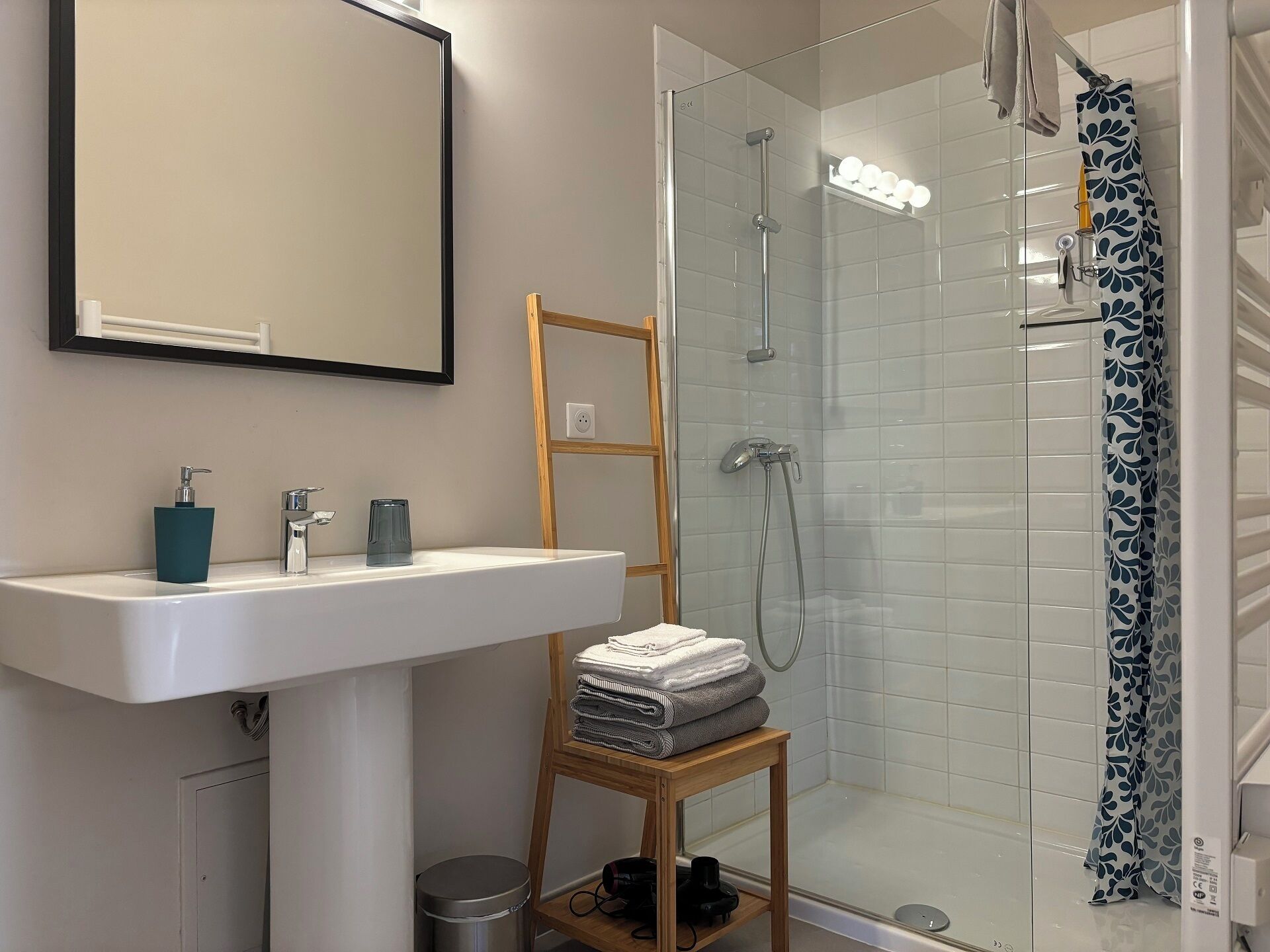 Un baño moderno con un lavamanos blanco, una cabina de ducha con paredes de vidrio y un estante de madera con toallas.
