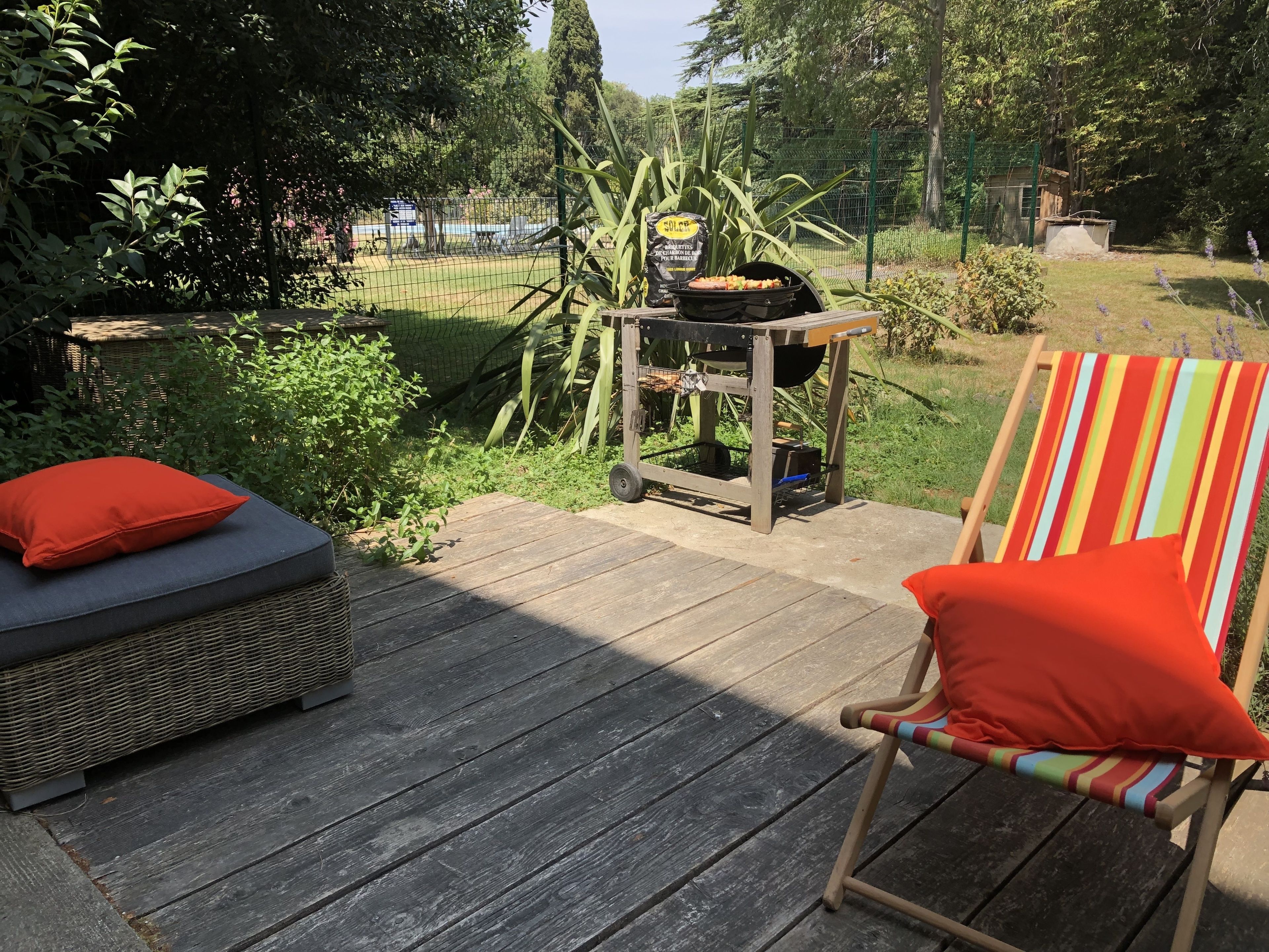 A wooden deck with a wicker sofa and a striped chair, a grill, and a garden in the background.