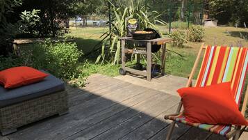 A wooden deck with a wicker sofa and a striped chair, a grill, and a garden in the background.