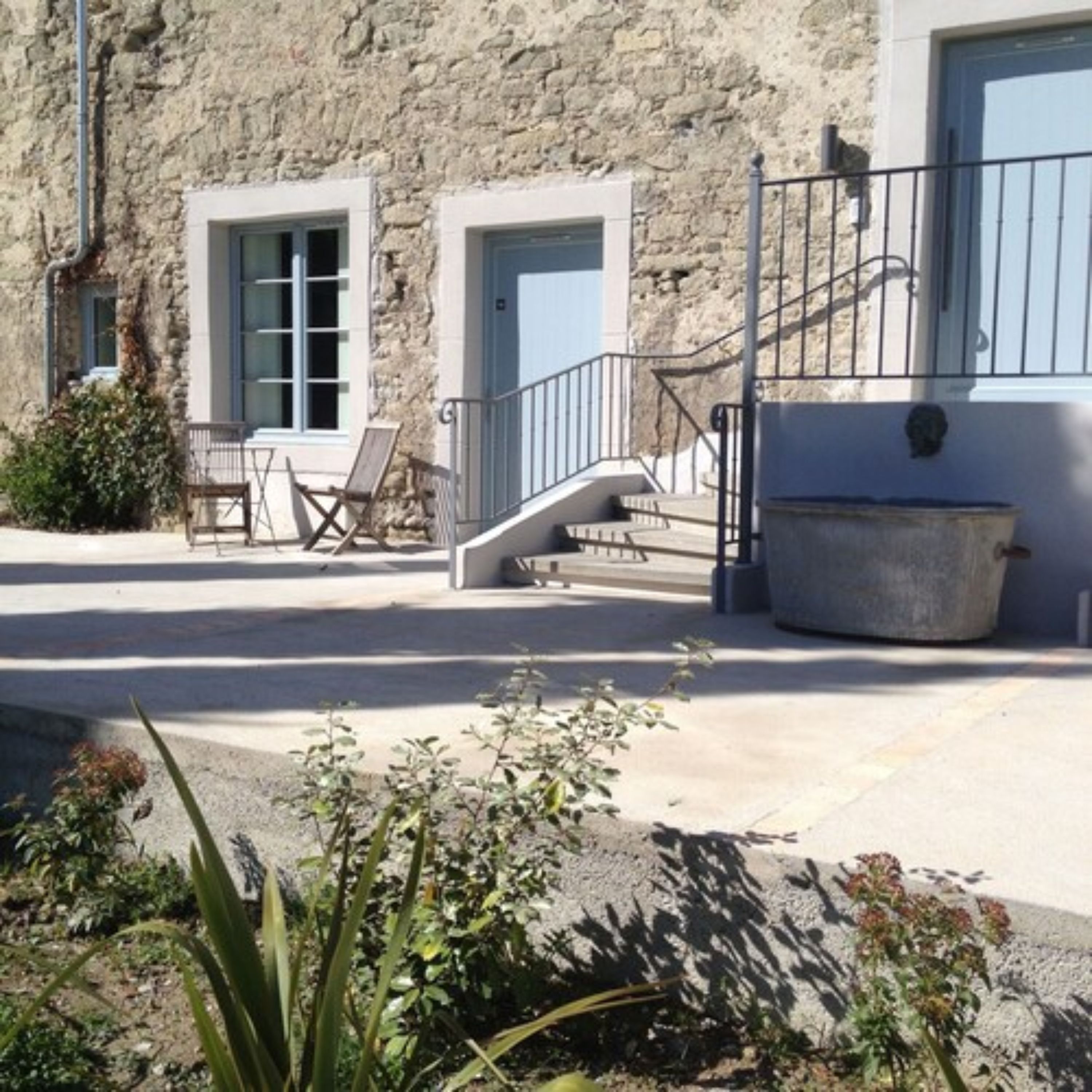 A stone building with two windows and a door, a staircase, outdoor seating, and a planter.
