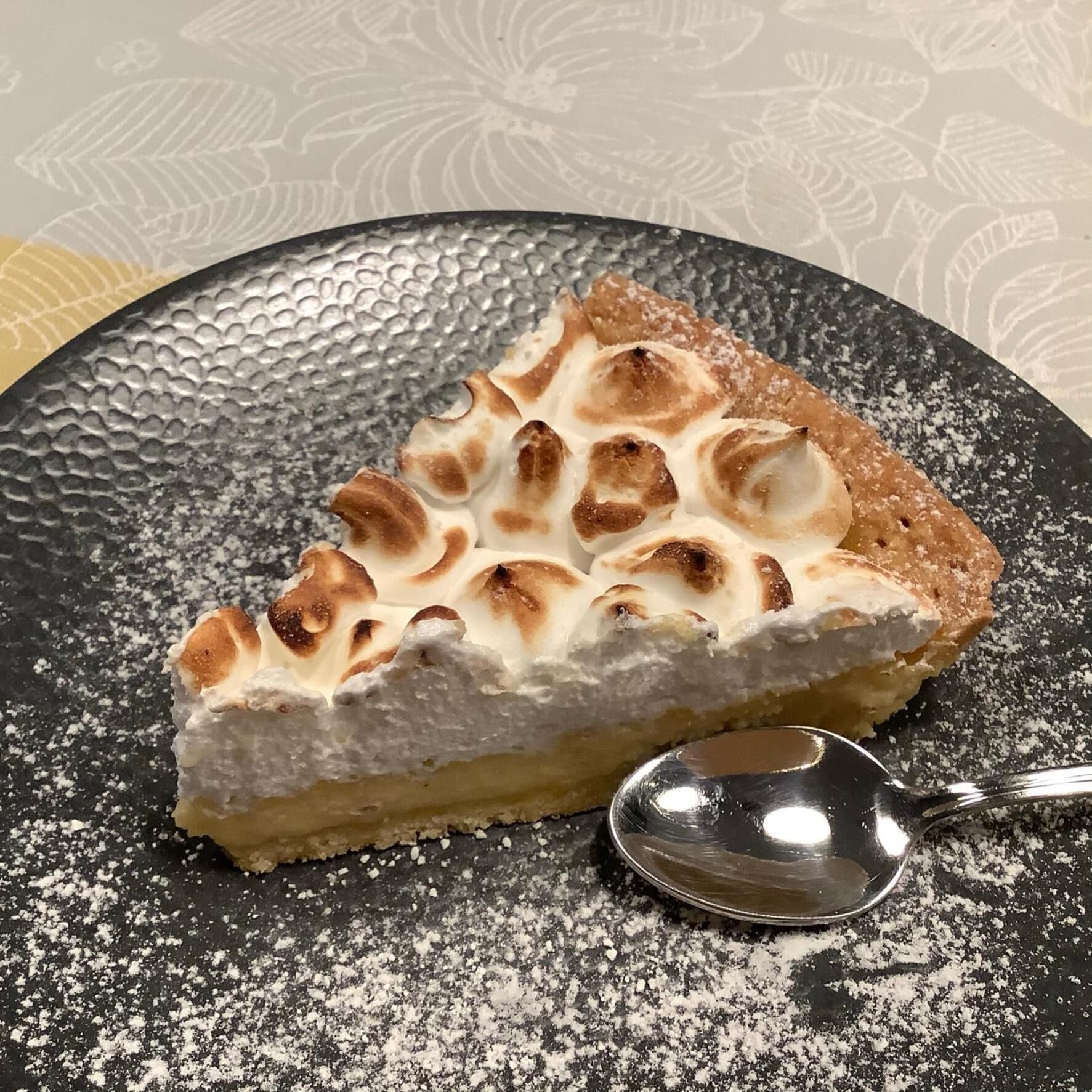 Une part de tarte à la croûte dorée et à la garniture crémeuse, saupoudrée de sucre glace, présentée sur une assiette à motifs.