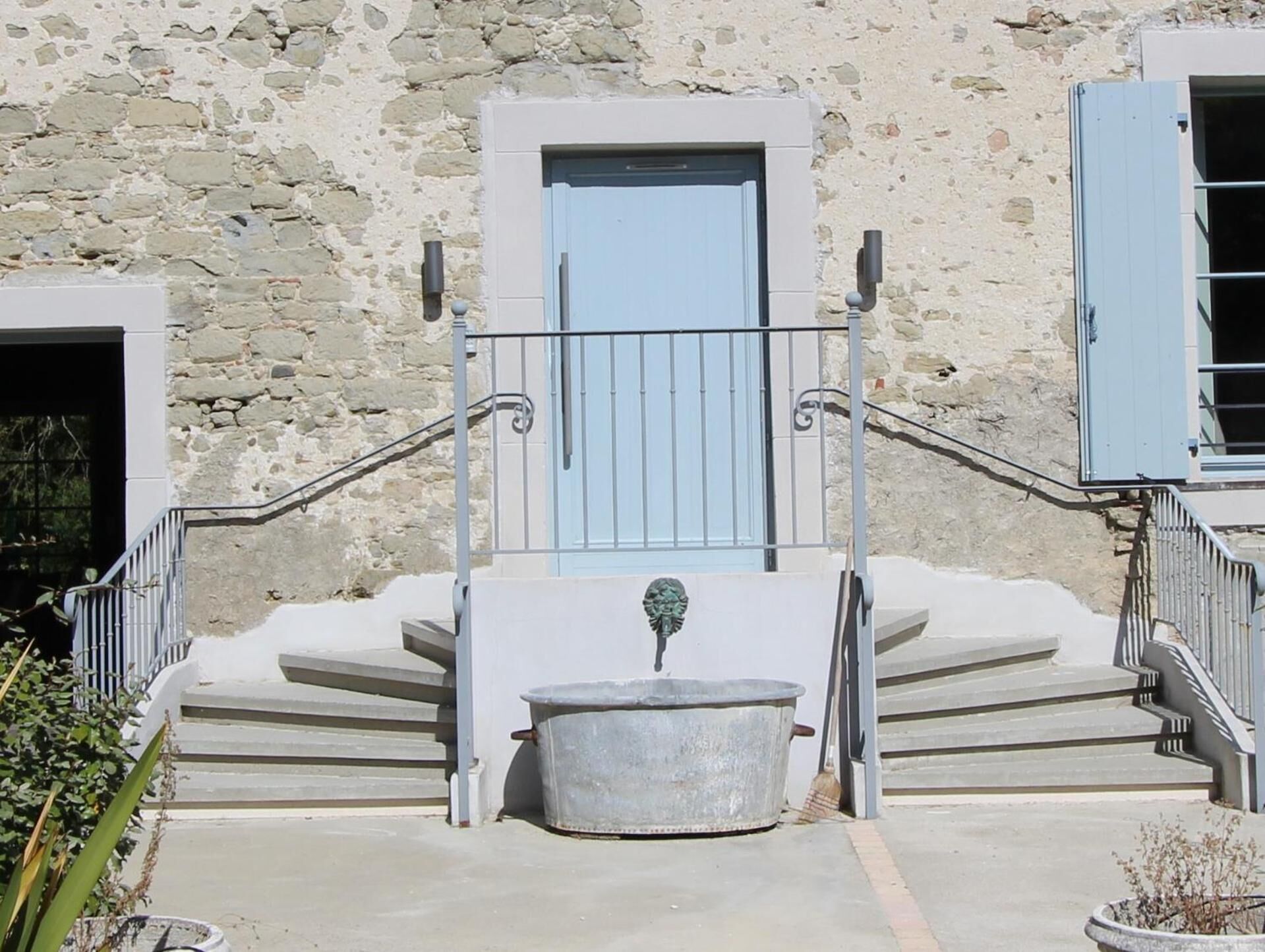 Un bâtiment en pierre avec une porte bleu clair, un escalier en métal doté d’une balustrade et un grand bac en métal devant l’entrée.