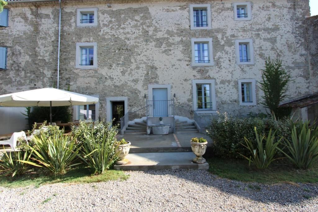 Un bâtiment rustique en pierre, doté de nombreuses fenêtres et d’une porte, entouré d’un chemin de gravier et de plantes en pot.