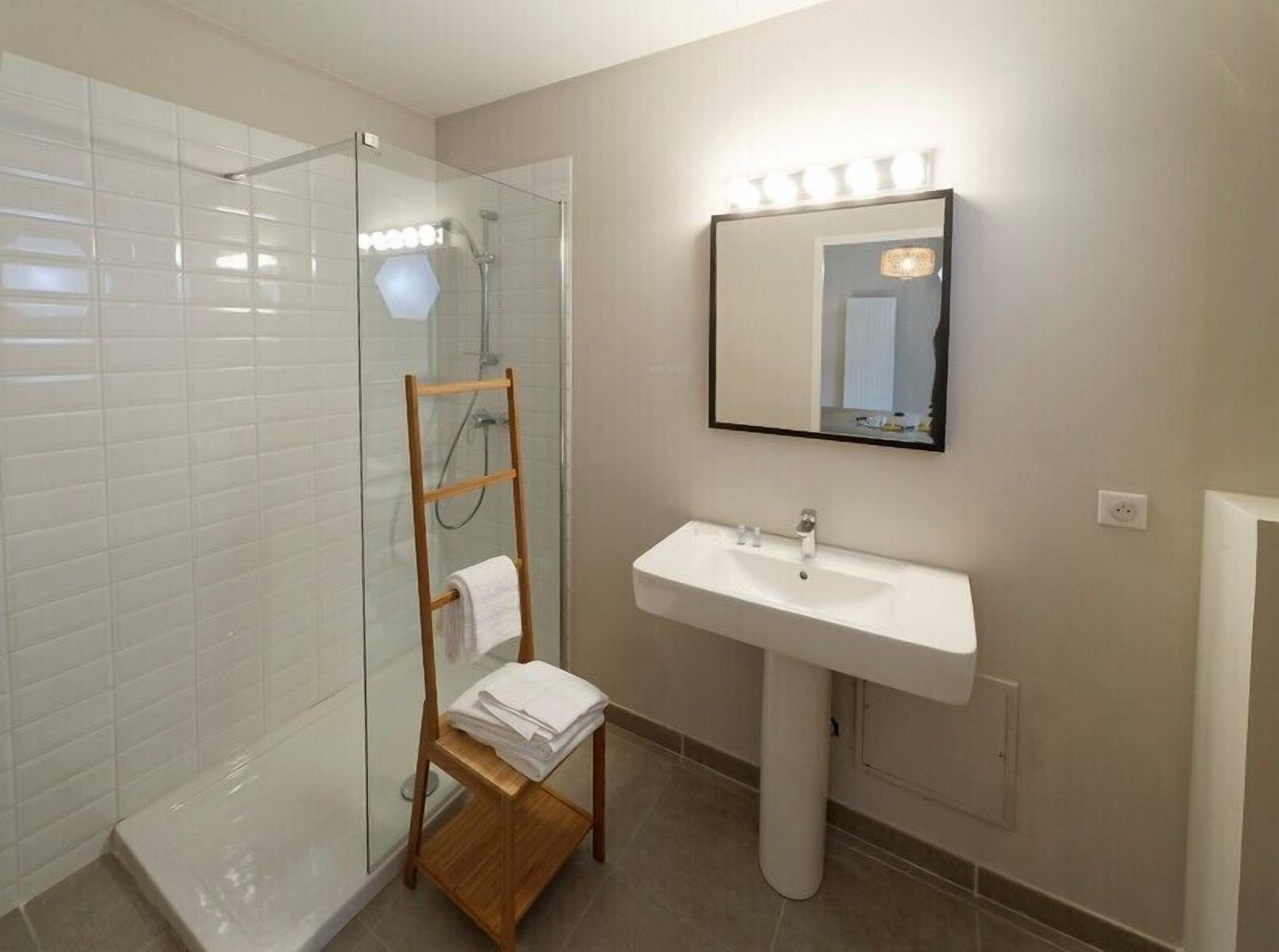 A modern bathroom with a glass shower enclosure, a wooden towel rack, a white sink, and a mirror with lights.