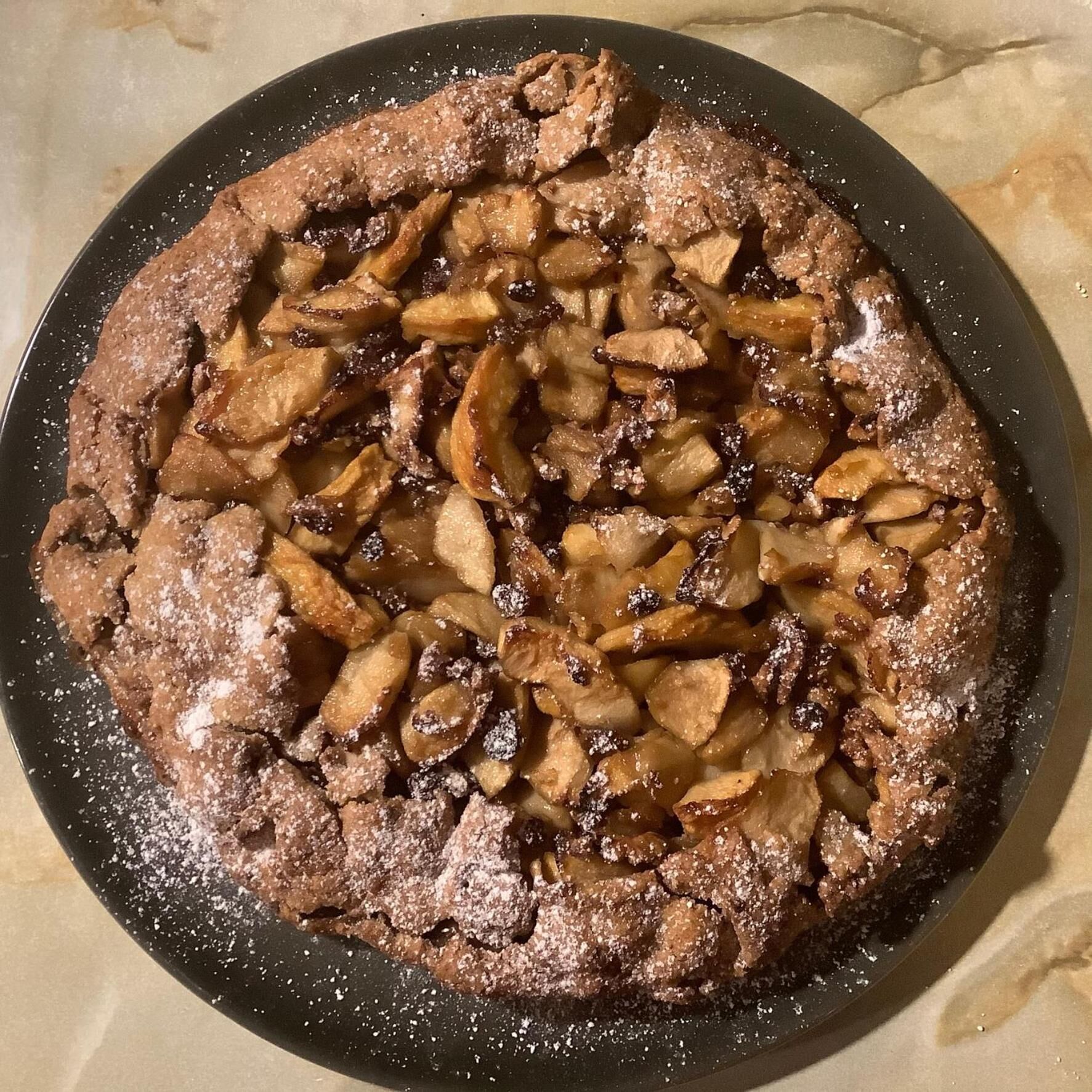 Un postre redondo con fruta en rodajas por encima, espolvoreado con azúcar glas, sobre un plato oscuro.